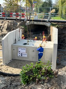 Een betonnen behuizing is met hijskettingen op de grond geplaatst. Vier bouwvakkers maken het stuk vast aan een ander betonnen behuizing.